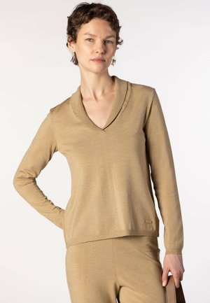 Femme aux cheveux courts et bouclés portant un haut beige à manches longues et un pantalon assorti, tenant un sac à main beige sur un fond clair.
