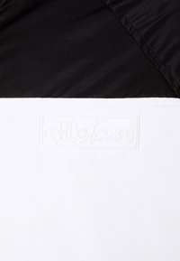 Schwarz-weiße Halbzip-Jacke mit glatter Textur, featuring ein erhabenes weißes Logo im unteren weißen Bereich und satinartigem Stoff.