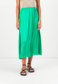 Fustă midi pliată verde, confecționată dintr-un material ușor, cu un brâu elastic. Asortată cu sandale negre cu bretele. Fundal alb simplu.