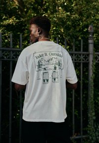 T-shirt couleur crème avec un motif au dos représentant des montagnes et deux chaises de camping, texte en vert : "Take It Outside".