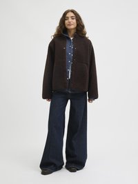 Pruun fliisjakker suurte taskute ja ees klõpsuga nööbiga, kantud teksapusero ja koos laia säärega tumedate teksapükstega.