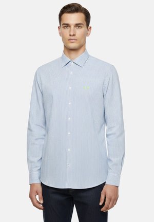 Uomo che indossa una camicia a righe sottili azzurro chiaro e bianca con bottoni e maniche lunghe, e pantaloni scuri, in piedi fermo davanti a uno sfondo semplice.