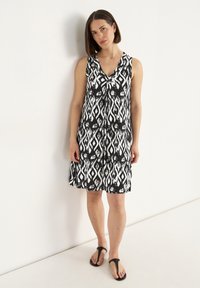 Ärmelloses Schwarz-Weiß-Kleid mit V-Ausschnitt, das ein abstraktes Diamantmuster zeigt und eine lockere A-Linie hat. Getragen mit schwarzen Flip-Flops.