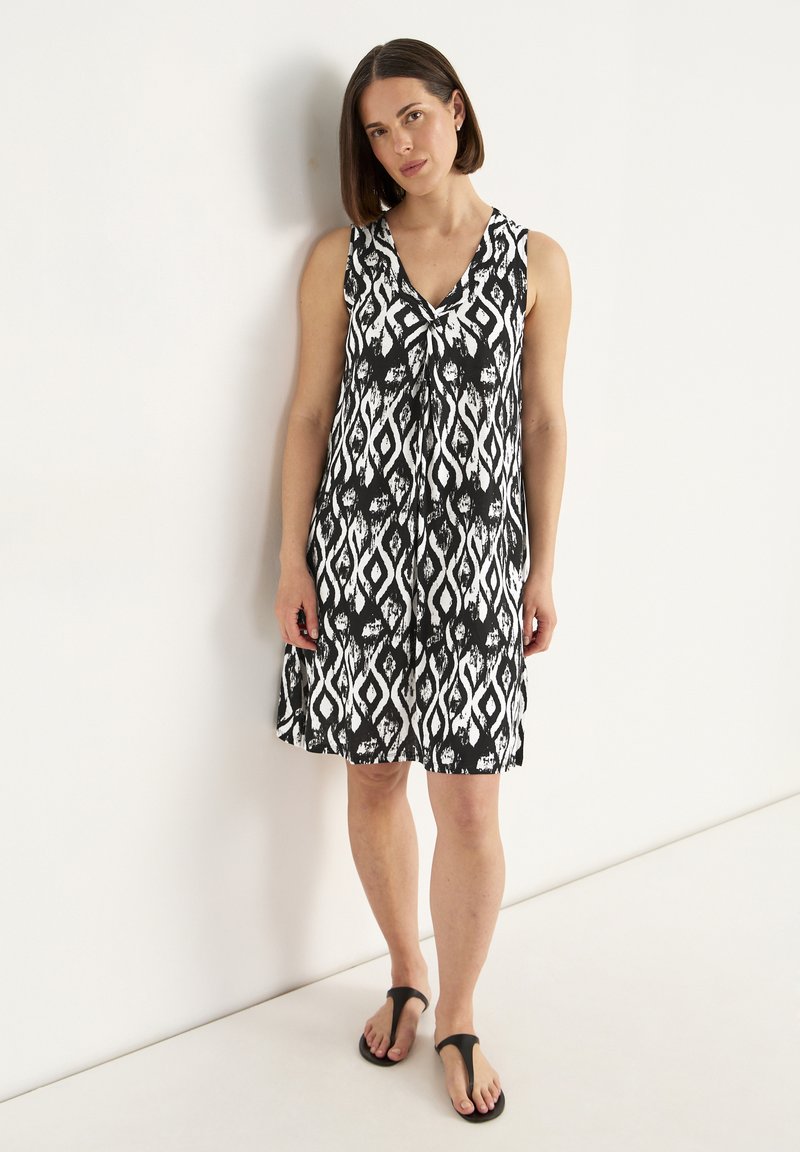 Ärmelloses Schwarz-Weiß-Kleid mit V-Ausschnitt, das ein abstraktes Diamantmuster zeigt und eine lockere A-Linie hat. Getragen mit schwarzen Flip-Flops.