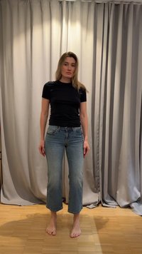 Γυναίκα που στέκεται ξυπόλυτη σε ξύλινο πάτωμα, φορώντας μαύρο μπλουζάκι και κοντά μπλε τζιν μπροστά από κλειστές μπεζ κουρτίνες.