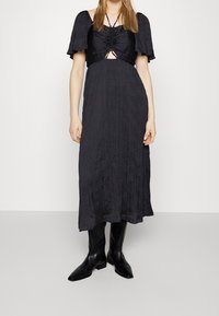 Vestido negro de media longitud con tejido texturizado, mangas cortas con vuelo y un corte clave atado en la cintura, combinado con botas negras.