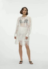 Vestido branco de renda com padrões recortados, mangas longas e um design transparente que termina acima do joelho. Usado com calçado preto.