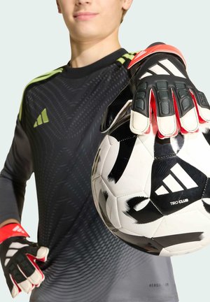 Joven portero de fútbol con camiseta negra sostiene un balón de fútbol Adidas Tiro Club con la mano enguantada extendida hacia la cámara.