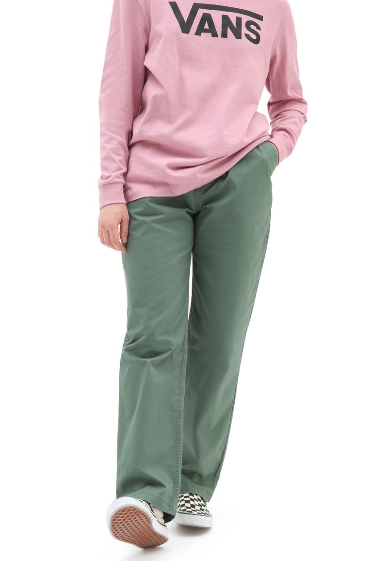 Pantalones de algodón verdes con un corte recto, combinados con una camiseta de manga larga rosa que presenta un logo negro. Zapatillas con un patrón en blanco y negro visibles.