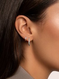 Gros plan sur l'oreille d'une personne aux cheveux lisses et foncés, portant une petite boucle d'oreille clou avec des pierres précieuses multicolores et une plus grande boucle d'oreille en argent.