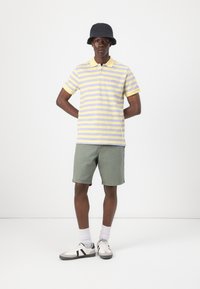 Stripet polo-shirt i gul og lavendel, parret med grønne shorts. En sort bucket hat og hvide sneakers med grå accenter fuldender outfittet.