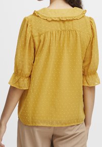 Gelbe Rüschbluse mit einem strukturierten Stoff mit kleinen Mustern, dreiviertellangen Puffärmeln und einem Rückenpasse-Design.