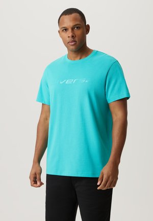 Turkusowa bawełniana koszulka z krótkim rękawem, okrągłym dekoltem i odblaskowym logo "diverse" na środku. Model w czarnych spodniach.