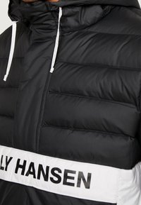 Giacca a vento nera con una banda orizzontale bianca con la scritta "HANSEN" in grassetto nero. Include un cappuccio e lacci bianchi a contrasto.