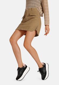 Gonna mini in denim khaki con spacco laterale, tasche frontali e un design aderente, abbinata a sneaker nere e rosa con una spessa suola bianca.