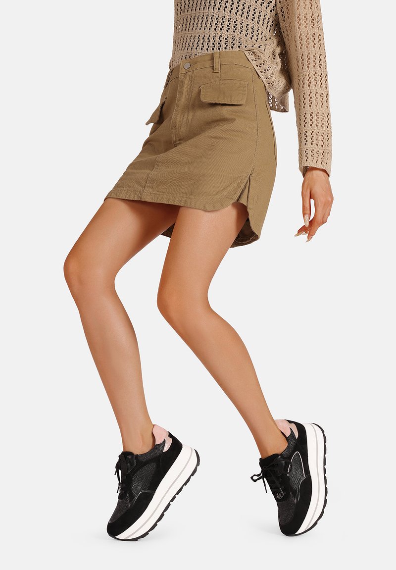 Gonna mini in denim khaki con spacco laterale, tasche frontali e un design aderente, abbinata a sneaker nere e rosa con una spessa suola bianca.