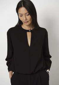 Schwarze Bluse mit langen Ärmeln, ausgestattet mit einem Schlitz-Ausschnitt und metallischen Akzenten. Glatter Stoff mit lockerer Passform, in taillierte Hosen gesteckt.