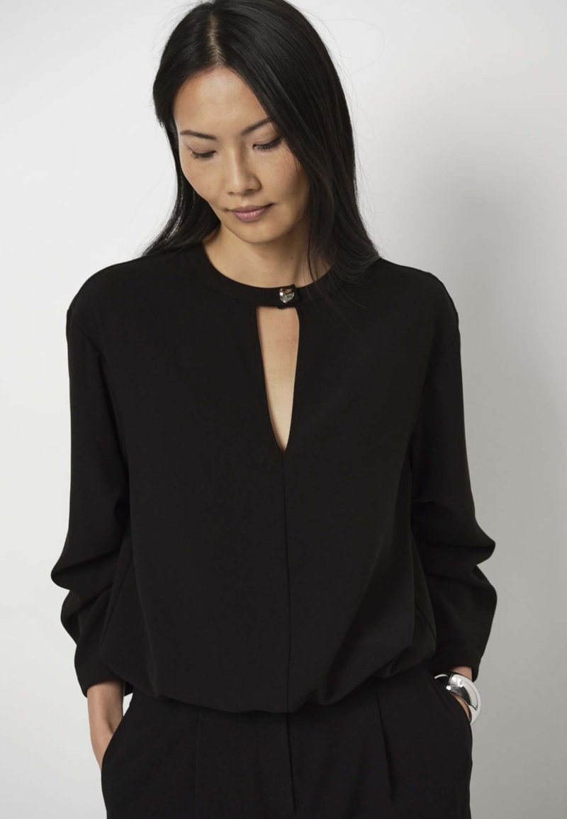 Schwarze Bluse mit langen Ärmeln, ausgestattet mit einem Schlitz-Ausschnitt und metallischen Akzenten. Glatter Stoff mit lockerer Passform, in taillierte Hosen gesteckt.