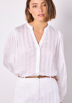 Weiße Bluse mit langen Ärmeln und vertikalen Falten, Knopfleiste und V-Ausschnitt. Kombiniert mit einem braunen Ledergürtel. Glattes, leichtes Material.