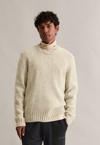 Sweater creme texturizado com um decote e bainha canelados, usado sobre uma blusa de gola alta bege clara, com mangas longas e um corte relaxado.