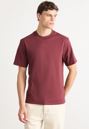 Jonge man in een effen bordeauxrode T-shirt met korte mouwen en lichtbeige broek, staande tegen een witte achtergrond.
