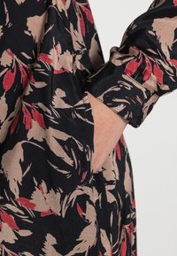 Sukienka w kwiatowy wzór w kolorach czarnym, beżowym i czerwonym. Długie rękawy zakończone mankietem. Widoczna kieszeń boczna.