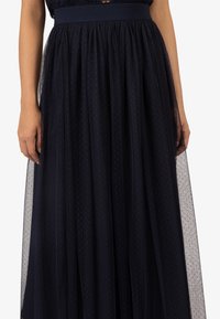 Marineblauwe tule rok met een geplooide tailleband. Heeft een gestructureerd patroon van kleine stippen en een vloeiend, gelaagd ontwerp voor extra volume.