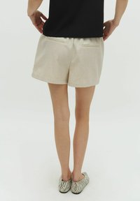 Pantaloni beige con due tasche posteriori, realizzati in un tessuto testurizzato. L'orlo è arrotolato e il modello indossa delle ballerine con fantasia.