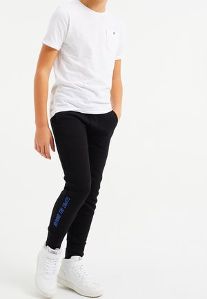 Zwarte trainingsbroek met blauwe tekst "BEYOND THE LIMITS" op het linkerbeen, gecombineerd met een wit T-shirt met ronde hals en witte sneakers.