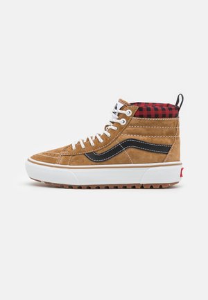 Sneaker alta in suede marrone con lacci bianchi, striscia nera, suola bianca spessa e colletto imbottito a scacchi rossi e neri su sfondo bianco.