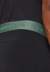 Juodieji sportiniai šortai su tekstūruotu žaliu diržu, kuriame tamsiai žaliais rašmenimis užrašyta "THE NORTH FACE". Tinkamas dizainas ir minkšta medžiaga.
