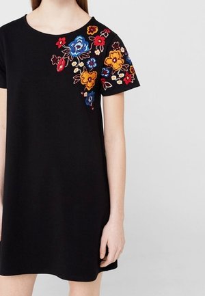 Robe noire à manches courtes avec des fleurs brodées colorées en rouge, bleu et jaune sur l'épaule gauche et la manche, portée par une personne debout.