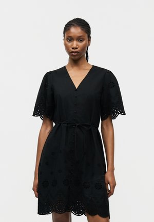 Kobieta z warkoczami, ubrana w czarną sukienkę na guziki z falbankowymi, ażurowymi koronkowymi rękawami i dołem, przewiązaną w talii cienkim paskiem.