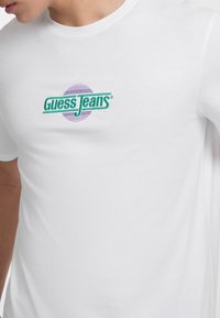 Balta medvilninė marškinė su daugiaspalviu siuvinėtu "Guess Jeans" logotipu ir apvaliu violetiniu akcentu. Klasikinis apvalios kaklo dizainas.