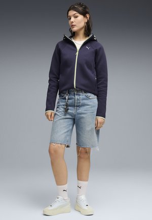 Jonge vrouw die een marineblauwe hoodie met rits draagt, lichtblauwe denim shorts, witte Puma sokken en witte sneakers, staand tegen een grijze achtergrond.