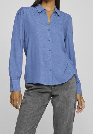 Kobieta ubrana w niebieską koszulę na guziki z delikatnym wzorem w kropki oraz szare dżinsy, stojąca z jedną ręką opuszczoną wzdłuż ciała.