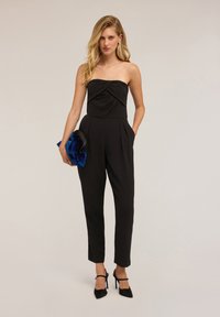 Svart strapless jumpsuit med en vriden bodice, smala ben och sidofickor, matchad med en blå satin-clutch och svarta klackar.