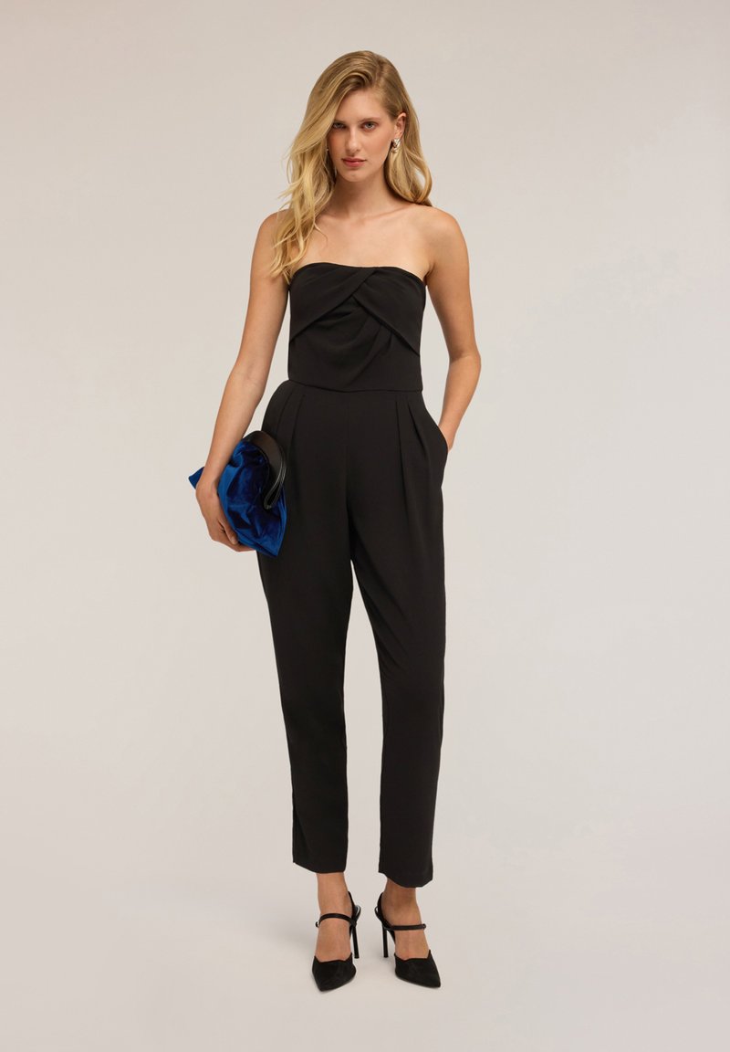 Svart strapless jumpsuit med en vriden bodice, smala ben och sidofickor, matchad med en blå satin-clutch och svarta klackar.