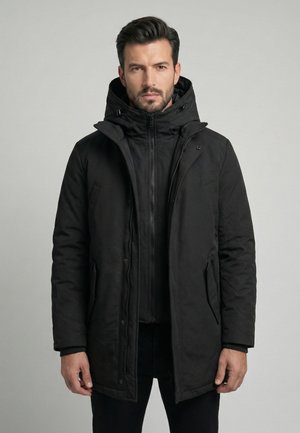 Homme portant un manteau d'hiver noir à capuche par-dessus une veste zippée, debout devant un fond gris clair uni.