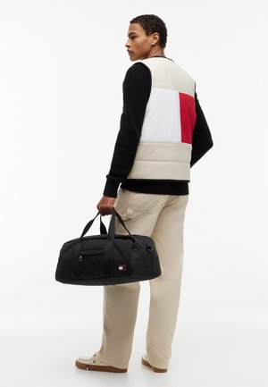 Persona joven vistiendo un chaleco con bloques de colores y pantalones beige, sosteniendo una bolsa de lona negra con un pequeño logo, de perfil contra un fondo blanco.