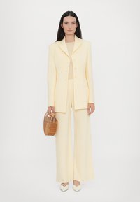 Light yellow tailored suit with a structured blazer, wide-leg pants, and a fitted top. Model holds a woven brown bag, wearing pointed white heels.