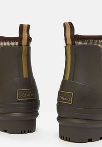 Barna gumis boka csizma textúrált felülettel, kockás bélésse és arany kiegészítőkkel, beleértve egy logó címkét és csíkos részleteket.