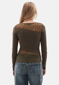Olijfgroene top met lange mouwen, voorzien van doorzichtige kantpanelen op de schouders en de onderrug. Gepaarde met hoge taille blauwe jeans.