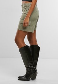 Oliwkowo zielone szorty cargo z dwoma przednimi kieszeniami, zestawione z wysokimi czarnymi skórzanymi botkami na obcasie. Gładka faktura i dopasowany krój.