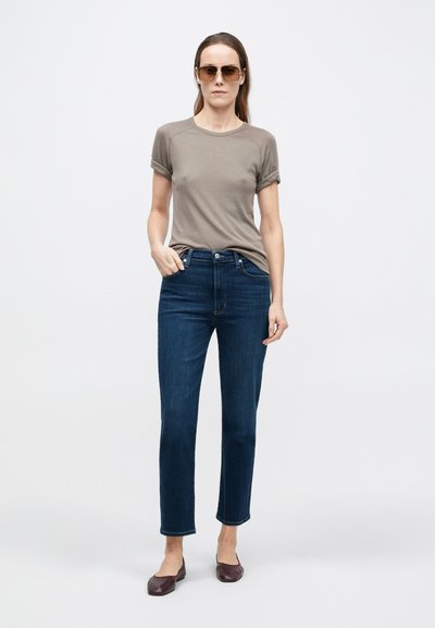 Világosbarna rövid ujjú póló felhajtott mandzsettákkal, sötétkék egyenes szárú farmerral és barna balerinacipővel párosítva, napszemüveggel kiegészítve.