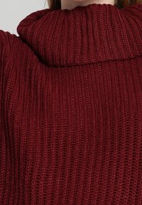 Bordowy sweter z dzianiny z wysokim, prążkowanym kołnierzem. Tkanina ma teksturowany wzór z pionowymi paskami.