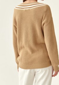 Beiger gestrickter Pullover mit lockerem Schnitt und kontrastierenden weißen Streifen am hinteren Kragen. Strukturierter Stoff und gerippte Bündchen.