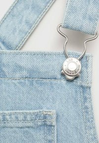 Svijetloplave traper kombinezone s metalnom kopčom i detaljima šivanja. Tkanina ima mekanu teksturu, a vidljiva je i prednja džep.