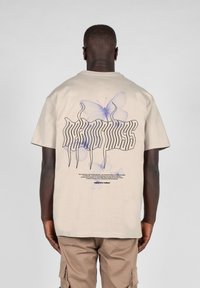 Beige bomulds t-shirt med et lilla sommerfugle grafik og stiliseret tekst på ryggen. Korte ærmer, afslappet fit og rund halsudskæring.