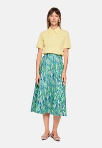 Poloshirt in hellem Gelb mit Kragen, kombiniert mit einem fließenden Midirock mit grün-blauem abstraktem Muster. Rosa Schuhe runden den Look ab.